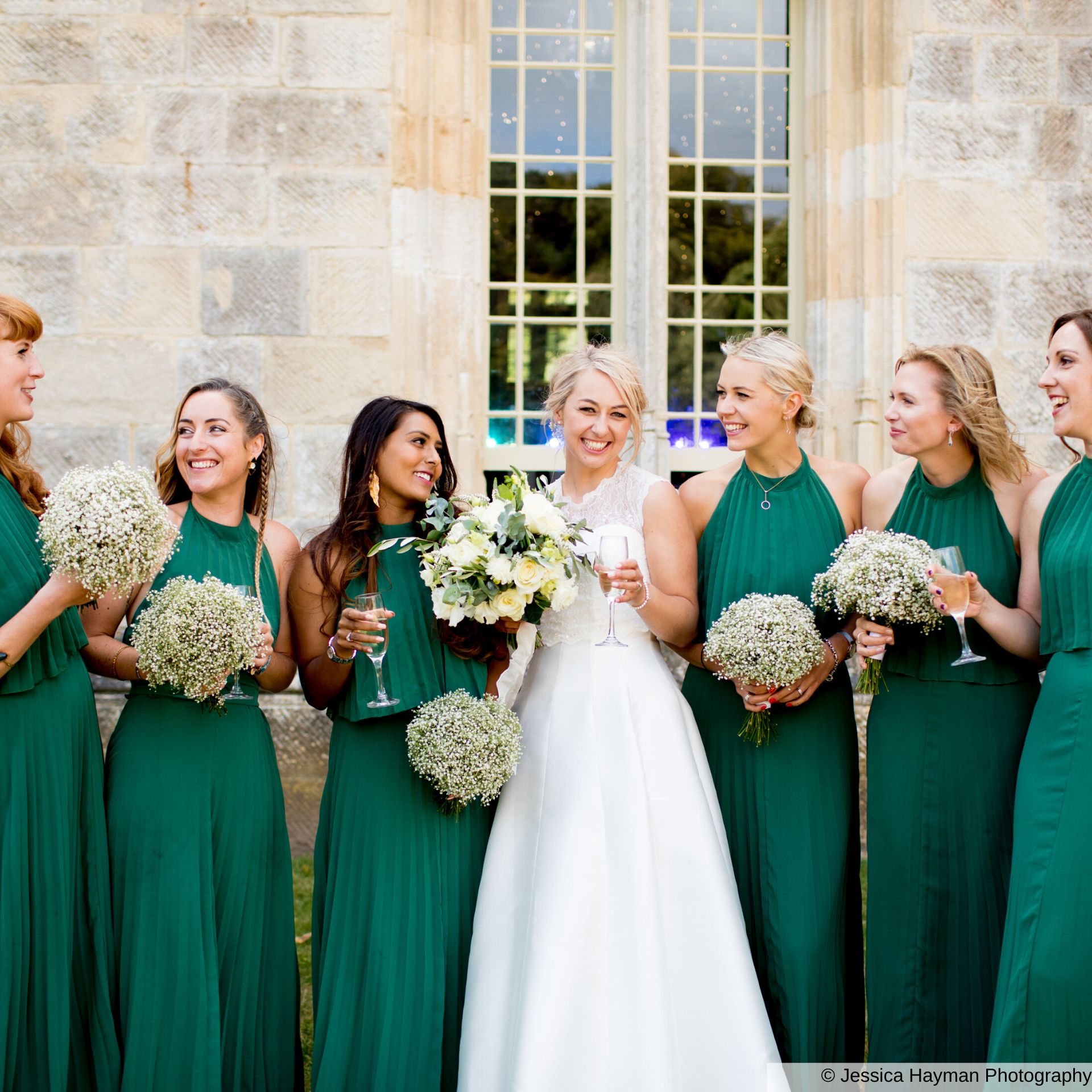 Bride and her bridesmaids dressed in emerald green, smiling and laughing in front of the beautiful stonework backdrop of Highcliffe Castle.