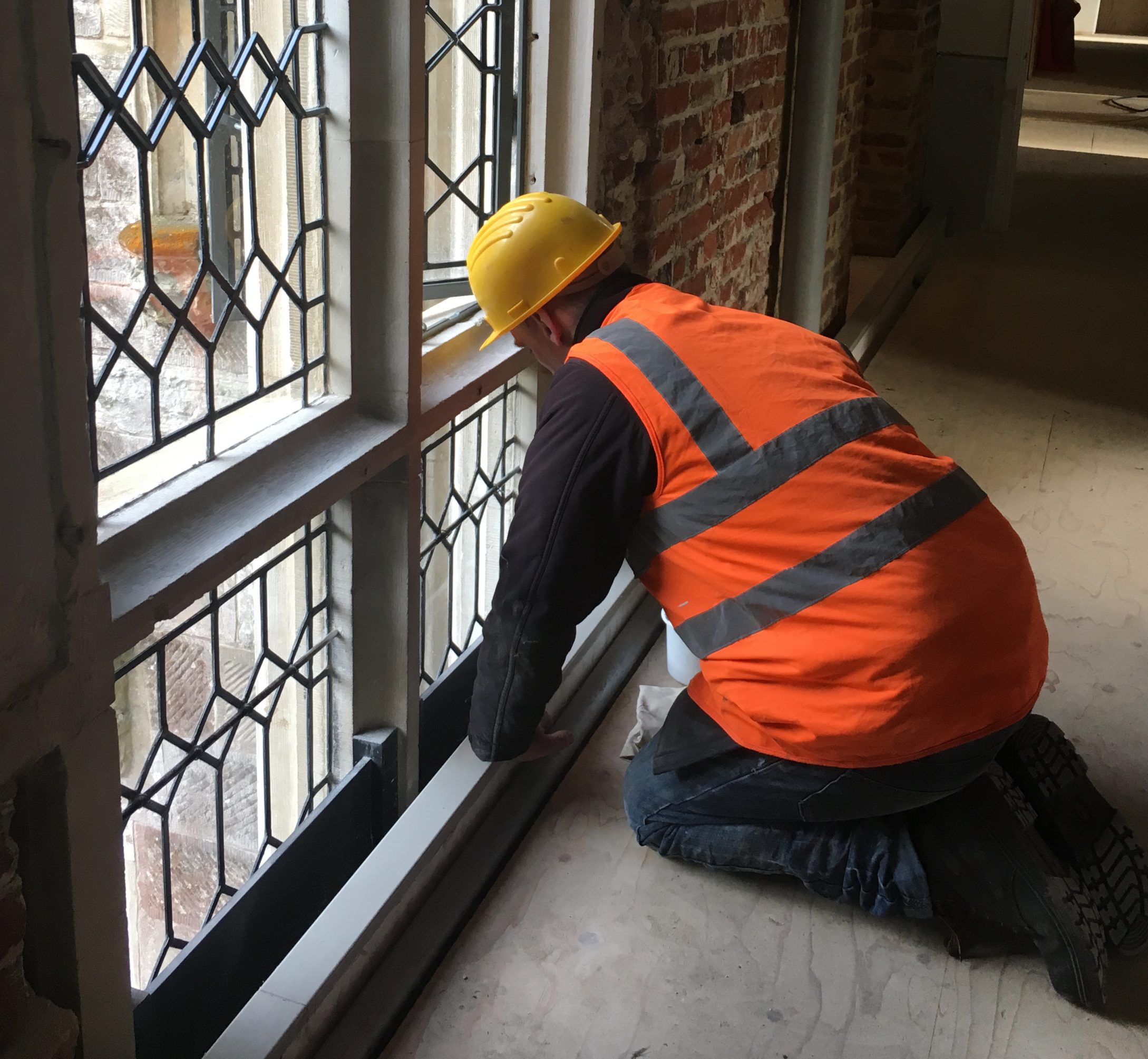 Man in high vis jacket and hard hat kneeling and working on repair to ornate mullion window with leaded panes.