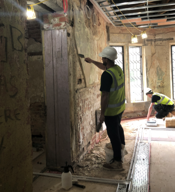 Workman in high viz vest brushing loose plaster from old brick wall in historic building