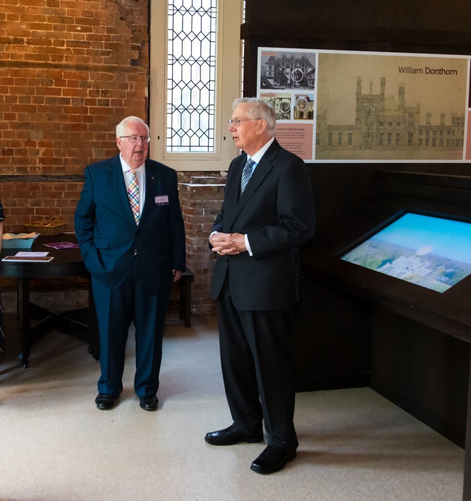 Two men in suits standing together talking with picture of Castle behind them.